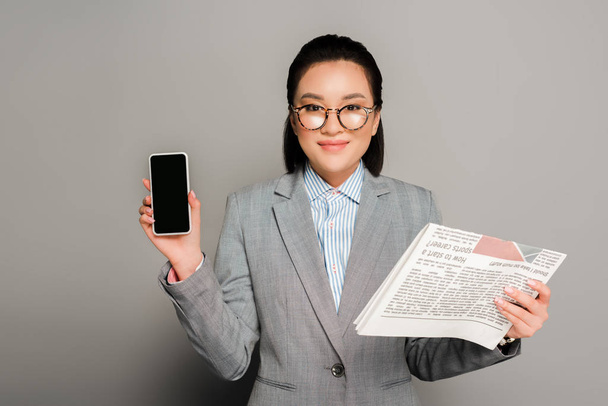 young businesswoman in eyeglasses holding newspaper and smartphone on grey background - Fotografie, Obrázek