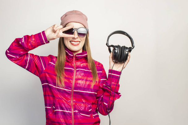 Friendly emotional. Young woman with glasses, a hat and a pink sports jacket holds headphones on a white background with copy space. Modern trendy music concept. Face expression. Banner. - Photo, image
