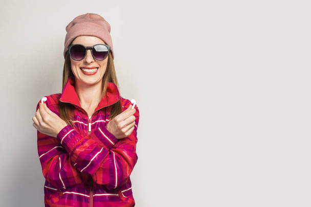 Přátelská Mladá žena v brýlích, klobouku a růžové sportovní bundě s usměvavou tváří drží bezdrátová sluchátka na bílém pozadí. Koncept moderního stylu, cool hudba. Výraz tváře. Nápis. - Fotografie, Obrázek