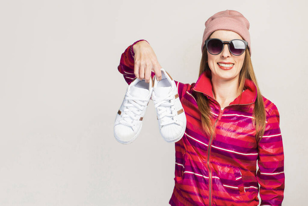 Дружеские эмоции. Молодая женщина в очках, шляпе и розовой спортивной куртке с улыбкой держит модные кроссовки на белом фоне с копировальным пространством. Современный стиль. Лицо. Баннер. - Фото, изображение
