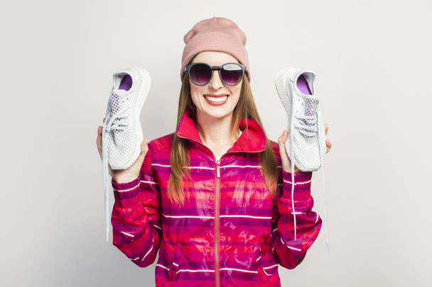 Emocionante amigável. Mulher jovem com óculos, chapéu e jaqueta de esportes rosa com um sorriso mantém tênis na moda em um fundo branco com espaço de cópia. Conceito estilo moderno. Expressão facial. Banner. - Foto, Imagem