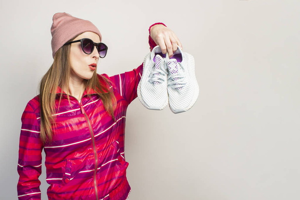 Vriendelijke emotionele Jonge vrouw in bril, hoed en roze sportjasje met een verrast gezicht houdt sneakers op een witte achtergrond met kopieerruimte. Concept moderne trendy stijl. Gezichtsuitdrukking. Banner. - Foto, afbeelding