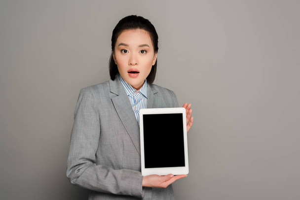 shocked young businesswoman in suit holding digital tablet on grey background - Fotoğraf, Görsel