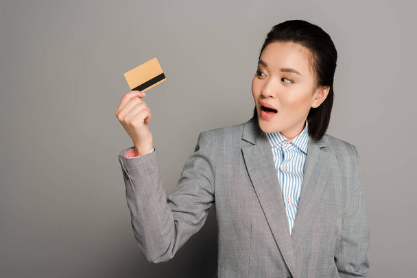 shocked young businesswoman in suit holding credit card on grey background - Foto, Imagem