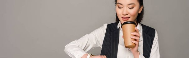 smiling young businesswoman looking at wristwatch and drinking coffee isolated on grey, panoramic shot - Fotó, kép