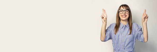 Χαμογελαστή νεαρή κοπέλα κρατά σταυρωμένα τα δάχτυλα σε ένα ελαφρύ φόντο. Έννοια του να κάνεις μια ευχή, καλοκαίρι. Μπάνερ. - Φωτογραφία, εικόνα