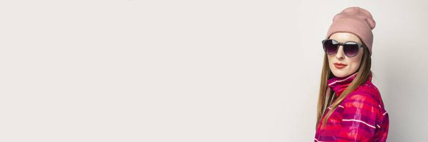 Amistosa mujer joven emocional con gafas, un sombrero y una chaqueta deportiva rosa con una cara seria sobre un fondo blanco con espacio para copiar. Concepto de estilo moderno de moda. Expresión facial. Banner
. - Foto, Imagen