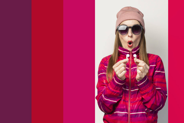 Emocionální Mladá žena v brýlích, klobouku a růžové sportovní bundě s překvapeným obličejem drží bezdrátová sluchátka na bílém pozadí. Koncept moderního stylu, cool hudba. Výraz tváře. Nápis. - Fotografie, Obrázek