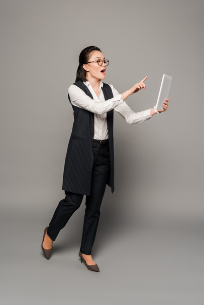 young businesswoman in eyeglasses with open mouth pointing with finger at digital tablet on grey background - Fotoğraf, Görsel