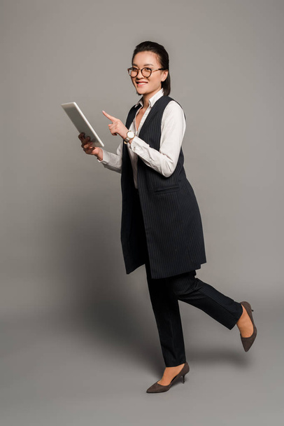 happy young businesswoman in eyeglasses pointing with finger at digital tablet on grey background - Foto, immagini