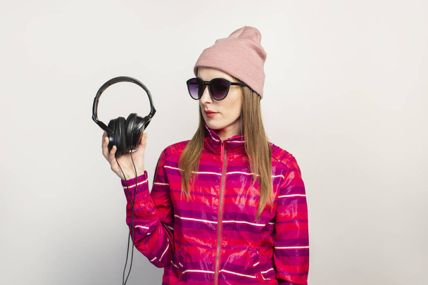 Friendly emotional. Young woman with glasses, a hat and a pink sports jacket holds headphones on a white background with copy space. Modern trendy music concept. Face expression. Banner. - Photo, Image