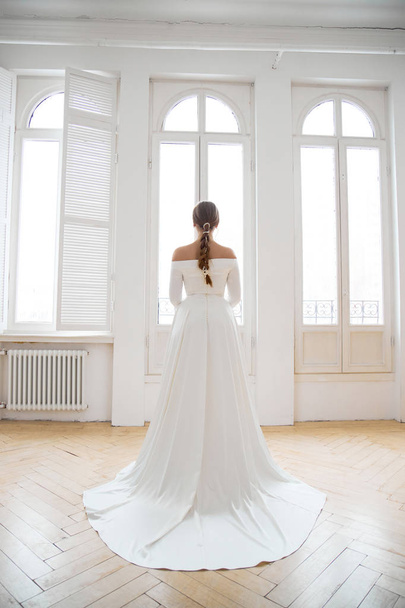 Beautiful slender blonde in the evening sun in a long white dress near a large window. Perfect hair and makeup for the bride, a new collection of wedding dresses - Photo, image