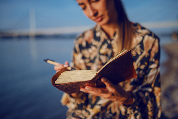 Portré elgondolkodtató vonzó kaukázusi barna, virágos ruha és fejpánt ül a sziklán a parton, a folyó mellett, és írás napló. - Fotó, kép