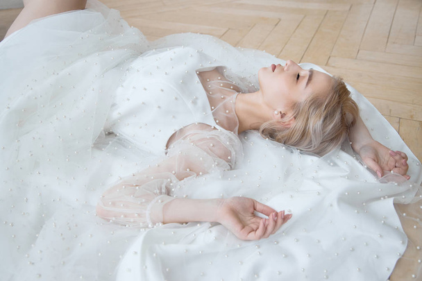Romantic portrait of a woman on the floor in a beautiful long white dress. The girl is blonde with blue eyes and beautiful makeup on her face. Natural cosmetics - Photo, Image