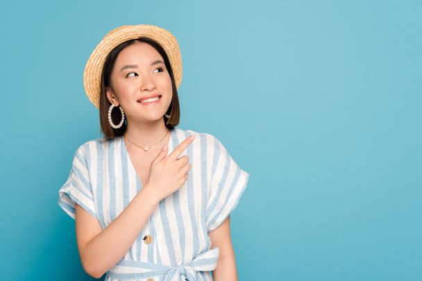 smiling brunette asian girl in striped dress and straw hat pointing with finger aside on blue background - Photo, Image