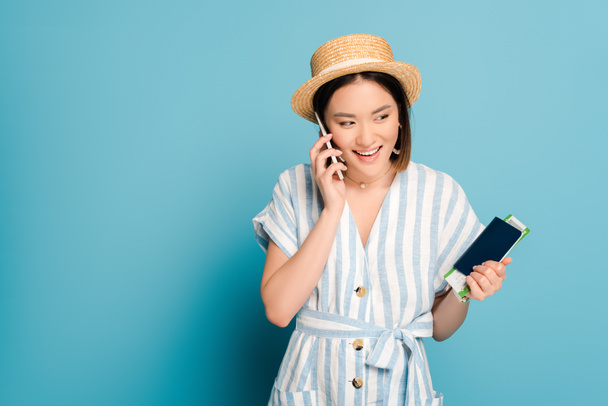 morena sonriente chica asiática en vestido a rayas y sombrero de paja con pasaporte con tarjeta de embarque mientras habla en el teléfono inteligente sobre fondo azul
 - Foto, Imagen