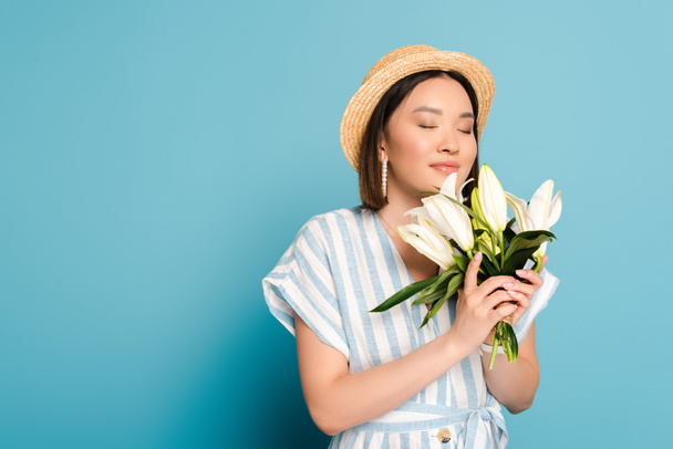 smiling brunette asian girl in striped dress and straw hat holding bouquet of lilies with closed eyes on blue background - Фото, зображення