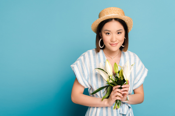 lächelnde brünette asiatische Mädchen in gestreiftem Kleid und Strohhut mit Lilienstrauß auf blauem Hintergrund - Foto, Bild