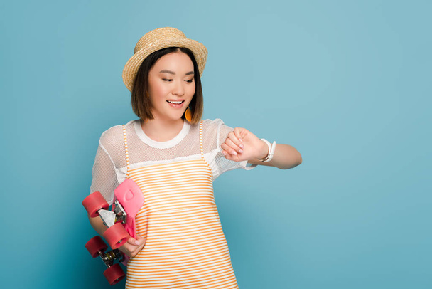 lachende aziatische meisje in gestreepte gele jurk en stro hoed met roze penny board kijken naar polshorloge op blauwe achtergrond - Foto, afbeelding