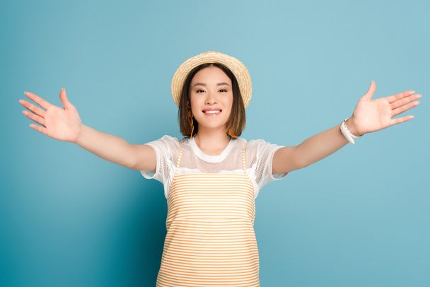 smiling asian girl in striped yellow dress and straw hat with open arms on blue background - Foto, Imagem