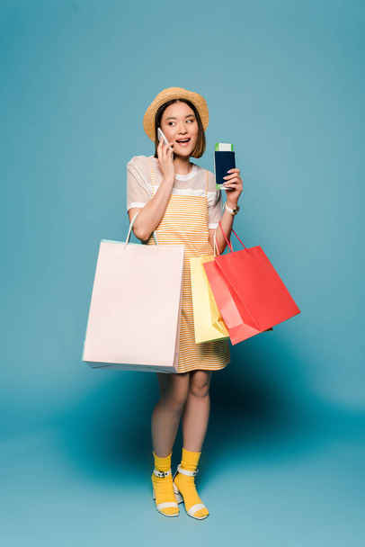 smiling asian girl in striped yellow dress and straw hat with shopping bags and passport talking on smartphone on blue background - Fotografie, Obrázek