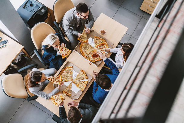 Pohled shora na hladové kolegy, kteří sedí u stolu a dávají si pizzu k obědu. Firemní pevný interiér. - Fotografie, Obrázek