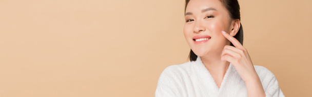 smiling beautiful asian woman in bathrobe touching face isolated on beige, panoramic shot - Fotoğraf, Görsel