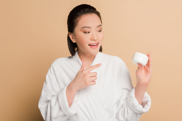 sorrindo bela mulher asiática em roupão apontando com o dedo para creme cosmético no fundo bege
 - Foto, Imagem