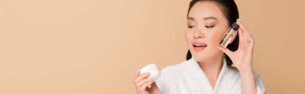 smiling beautiful asian woman in bathrobe holding cosmetic cream and oil isolated on beige, panoramic shot - Photo, Image
