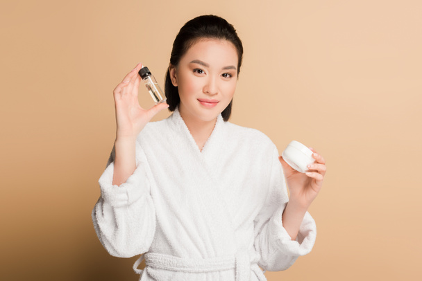 smiling beautiful asian woman in bathrobe holding cosmetic cream and oil on beige background - Фото, зображення