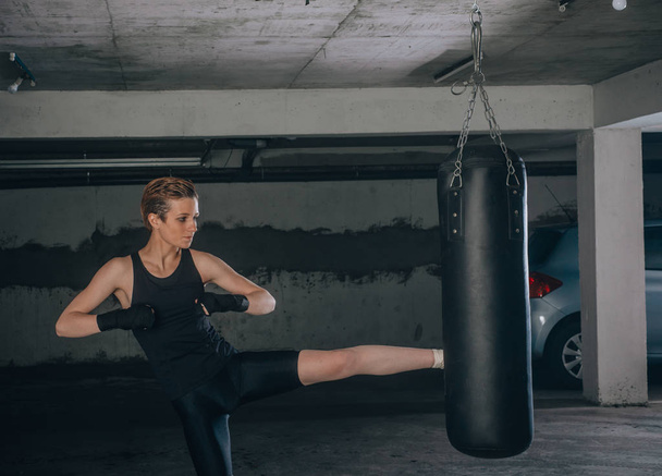 Biała kobieta w odzieży sportowej i z czarnymi bandażami kopiąca worek bokserski w garażu - Zdjęcie, obraz