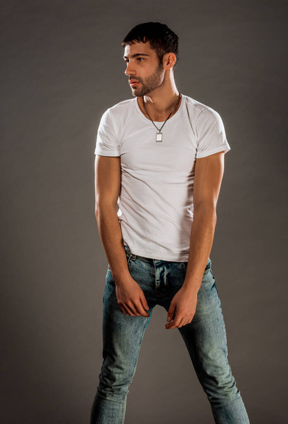 Aantrekkelijk jong mannelijk model poseren in blauwe jeans en witte top geïsoleerd op grijze muur - Foto, afbeelding