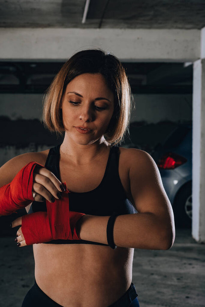 Closeup portrait of a beautiful woman doing red boxing bandages in a garage. - Фото, изображение