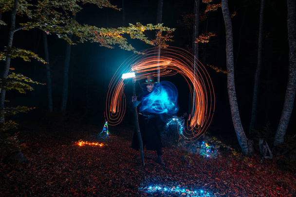 Hechicero (mago) lanzando un hechizo mientras está de pie en los bosques mágicos. Sostiene su bastón mágico. La iluminación mágica. Celebración de vacaciones de otoño. Concepto de misterio y pesadilla - Foto, Imagen
