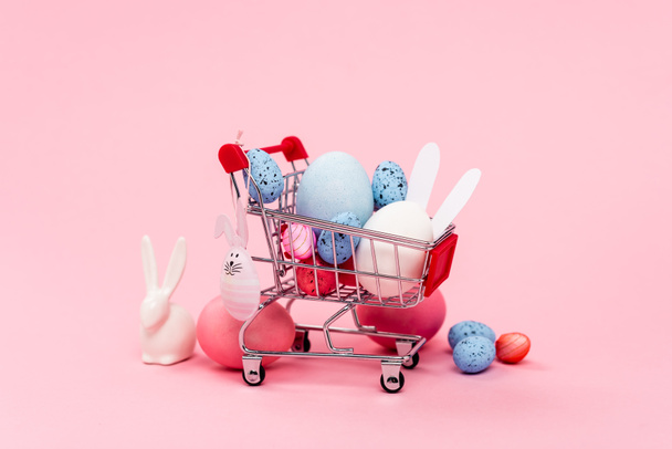 Bunte Ostereier und dekorative Hasen im Einkaufswagen auf rosa Hintergrund - Foto, Bild