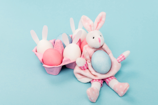 Pink egg tray with painted chicken eggs and decorative bunnies on blue, easter concept - Photo, Image