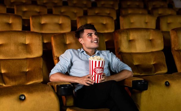 Um jovem sorrindo curtindo com suas pipocas enquanto assiste a um filme no cinema de teatro - Foto, Imagem