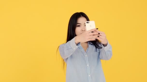  girl makes selfie Asian woman on yellow background admiring them through phone - Imágenes, Vídeo
