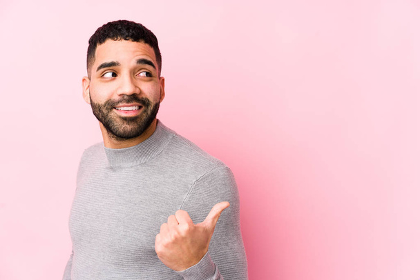 Junger lateinischer Mann vor rosa Hintergrund zeigt vereinzelt mit dem Daumenfinger weg, lacht und ist unbeschwert. - Foto, Bild