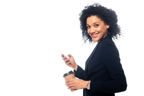 Widok z boku Afryki amerykańskiej kobieta uśmiecha się z jednorazowej filiżanki kawy i smartfona izolowane na białym - Zdjęcie, obraz