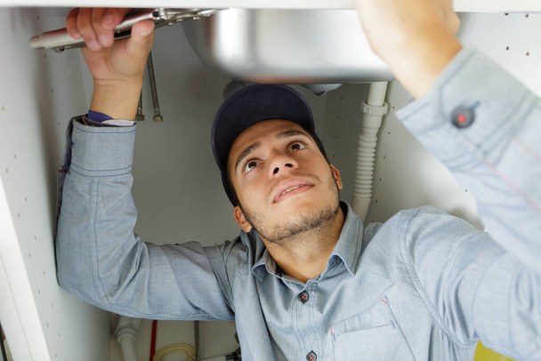 young male plumber performing a repair under the kitchen sink - Photo, Image