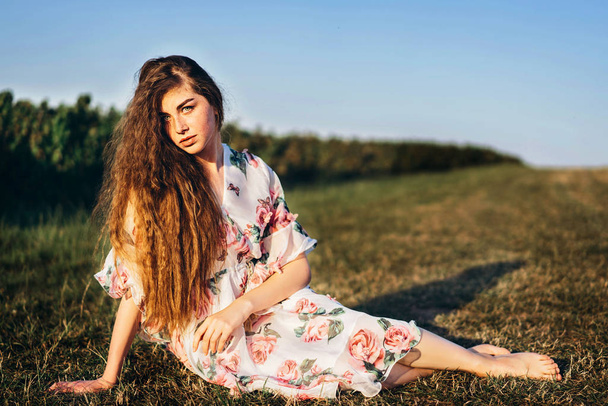 Full length portrait of beautiful woman with long curly hair on currant field background. Girl in a light dress sits on the grass in sunny day. - Foto, Imagen