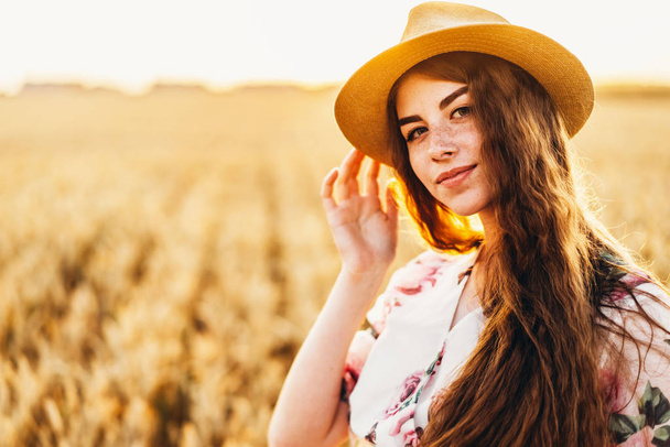 Portrait of a beautiful young woman with curly hair and freckles face. Woman in dress and hat posing in wheat field at sunset and looking at camera. - Foto, Imagen