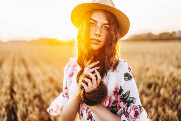 Portret van een mooie jonge vrouw met krullend haar en sproeten gezicht. Vrouw in jurk en hoed poseren in tarweveld bij zonsondergang en kijken naar camera. - Foto, afbeelding