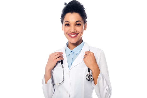 Front view of smiling and positive african american doctor with stethoscope looking at camera isolated on white - Foto, afbeelding