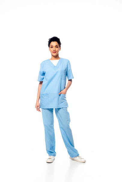 Full length view of african american nurse with hand in pocket smiling and looking at camera on white background - Photo, Image