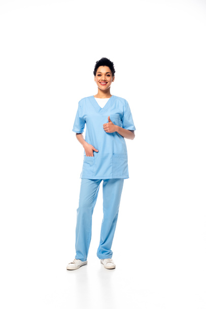 Дивлячись на медсестру-афроамериканку з рукою в кишені і великим пальцем, що посміхається і дивиться на камеру на білому - Фото, зображення
