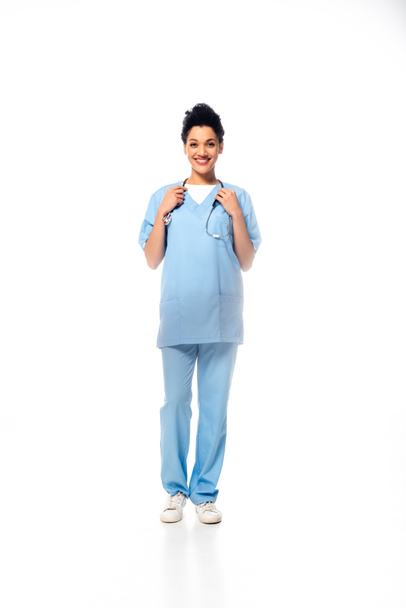Vollständige Ansicht der afrikanisch-amerikanischen Krankenschwester mit Stethoskop lächelnd auf weißem Hintergrund - Foto, Bild