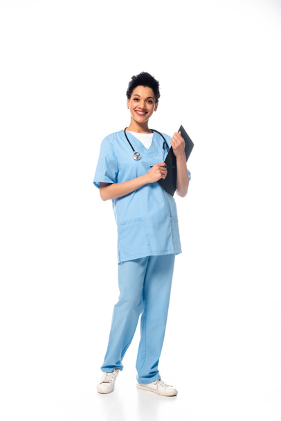 Full length view of african american nurse with stethoscope holding clipboard and smiling on white background - Fotografie, Obrázek