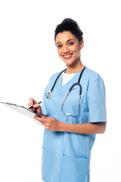African american nurse with stethoscope writing on clipboard and smiling isolated on white - Foto, Imagem